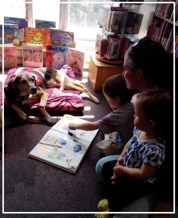 Image depicting a child seated next to her mother while reading to a service dog.